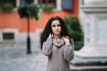 Pretty young woman dressed in gray sweater posing in the mediaeval city on a sunny day. Old town green garden and ancient windows background. Copy space
