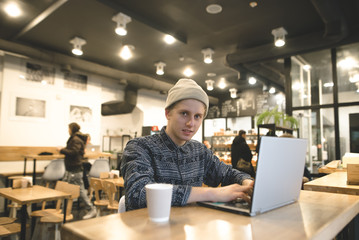 A hipster student sits in a cozy cafe with a laptop and works. A young man enjoys intranet on a...