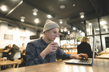 Fototapeta premium A young freelancer works for a laptop in a lush cafe and drinks coffee. Hipster student at a computer in a cafe.