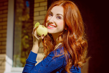 Beautiful cheerful woman eating a green apple, outdoors portrait, smiling woman with healthy teeth holding  apple. Very cute ethnic model with a red hair outdoors.
