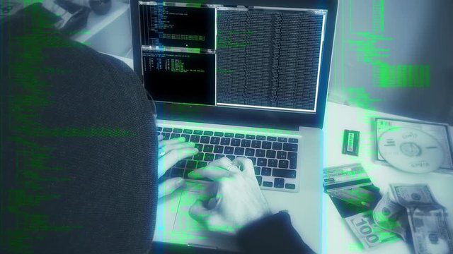 Hooded Hacker, Cyber Attack Crime Scene. Hooded person typing in a laptop next to stolen itens, simulating a hacker crime scene