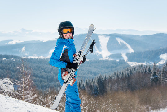 Happy female skier smiling to the camera, holding her skis, wearing blue ski suit and black helmet at winter ski resort copyspace happiness positivity vacation travelling
