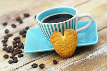 Crunchy cookie in shape of heart leaning against cup of coffee on rustic wooden surface
