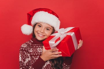 Portrait of pretty small adorable charming girl closing her eyes with cookies in shape of heart, wearing knitted traditional costume, x mas eve celebration, stand isolated on pure red background
