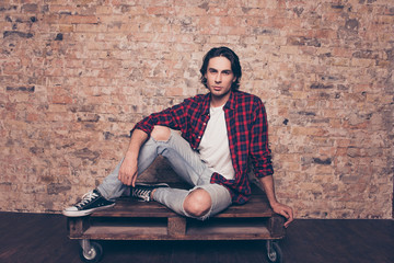 Full length close up of young successful freelancer student on the brick stones wall background, sitting on top of palette, in casual checkered outfit, jeans, sporty casual shoes