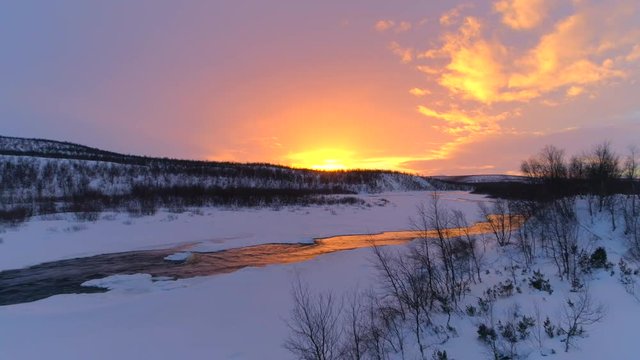AERIAL: Flying above icy meandering river flowing through snowy winter wonderland in Lapland wilderness at dreamy sunset. Stream meandering through frosty countryside on cold morning at golden sunrise