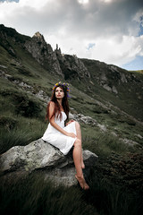 Naklejka na ściany i meble Beautiful woman in a long white dress in the mountains. Young woman sitting on a rock. Hair blowing in the wind. Rocky mountain. Beautiful woman with flower wreath