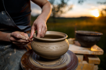 Clay potter creating on the pottery wheel. Sculptor from fresh wet clay on pottery wheel. Selected focus. Hands of young potter was produced on range of pot in open air - 180038841