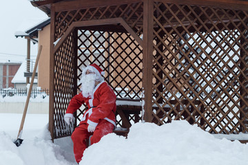 Tired santa claus sitting on wooden chair after work of snow removal in front of wooden house