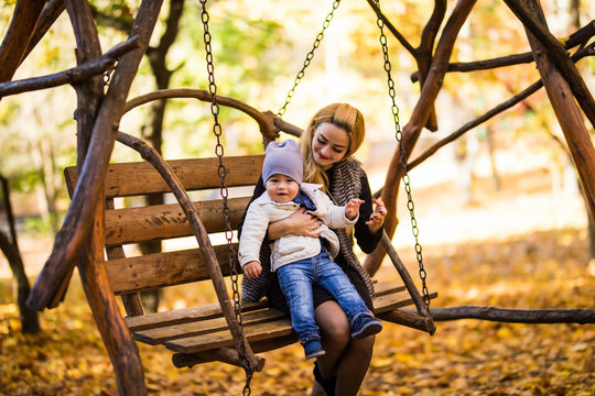 Young mother with her small son is resting on a wooden bench in the autumn golden park