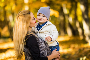 Mom and her son are in the autumn park, the son loves looking at his mother, the woman is holding his hands, in the background yellow leaves, the family spends the weekend in the park