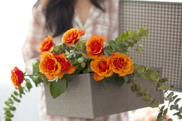 bouquet of orange roses in a gray box in female hands at home by the window