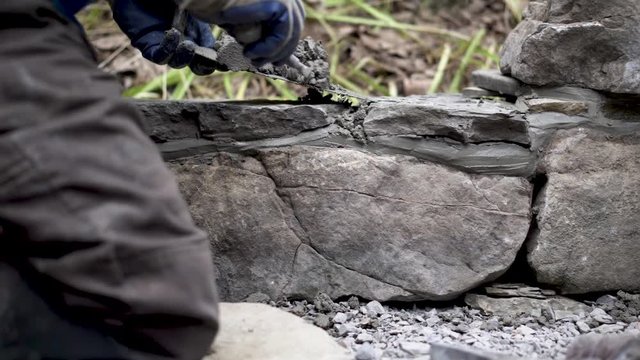 Using a trowel and a tuckpointing trowel a hardscape puts mortar between the stones in a rock wall.