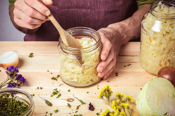 The cook puts sauerkraut in glass jars. On the table are spices, onions and cabbage. - obrazy, fototapety, plakaty