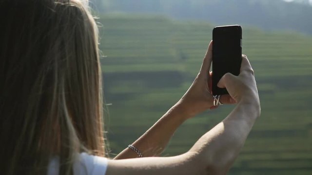 Young attractive woman watching sunrise over Longji Rice Terraces from top of the mountain. Blonde beautiful girl takes panorama photos on phone.