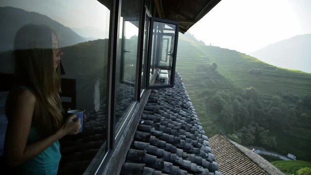 Young beautiful woman enjoys her morning tea looking at the mountains and rice fields in famous Ping An Village, Longji,China. Freedom,Calmness and relax concept.Attractive girl holding cup of coffee