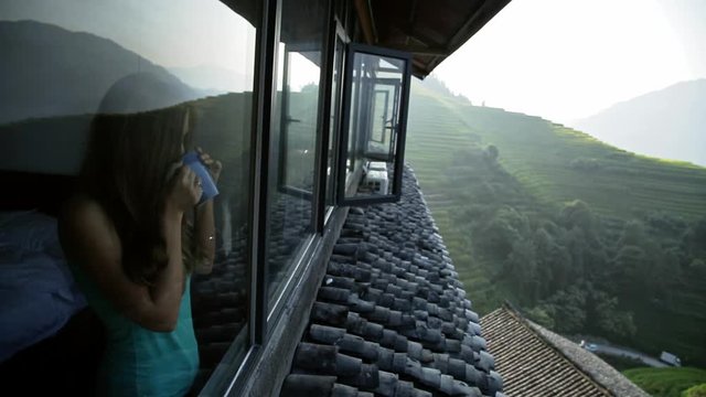 Young beautiful woman enjoys her morning tea looking at the mountains and rice fields in famous Ping An Village, Longji,China. Freedom,Calmness and relax concept.Attractive girl holding cup of coffee