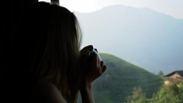 Young beautiful woman enjoys her morning tea looking at the mountains and rice fields in famous Ping An Village, Longji,China. Freedom,Calmness and relax concept.Attractive girl holding cup of coffee