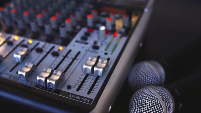 Close view of blurred sound console with many indicators, sliders, and buttos and closeup of two microphones