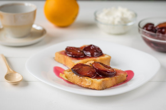 Toasted French bread, plum jam, cottage cheese and orange on a white wooden table.