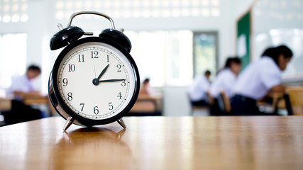 Retro alarm clock with one o'clock, left side on wood table teacher in blur students exams classroom. Time is indefinite continued progress of existence. Education exams concept, black and white - obrazy, fototapety, plakaty