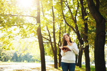 A beautiful happy smiling brown-haired woman in white sweater standing with a red book in fall city park on a warm day. Autumn golden leaves. Reading concept.