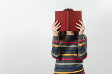 Beautiful European young brown-haired woman in glasses for sight dressed in casual dark grey longsleeve with colorful stripes, hiding behind book on a white background. Reading and studying concept.