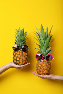 Female hands holding ripe pineapples with sunglasses on yellow background