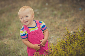 Happy cute baby girl with blond hairs wearing pink jeans dress posing smiling in central park between green tree and stone