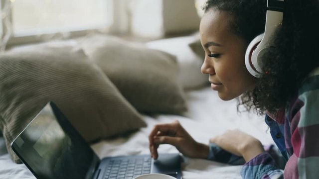 Close-up of african american girl listen to music while watching photos online on laptop lying on bed