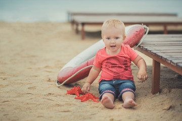 Happy cute baby girl with blond hairs and blue eyes wearing stylish clothes posing smiling on sea side beach on wooden way with safety round and red sea star