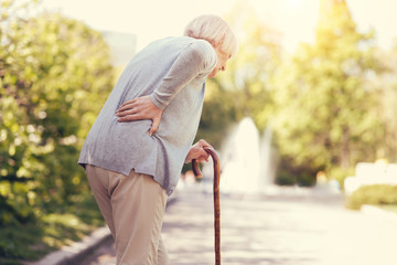 Old age. Sad aged unhappy woman holding a walking stick and walking while feeling pain in the back - Powered by Adobe