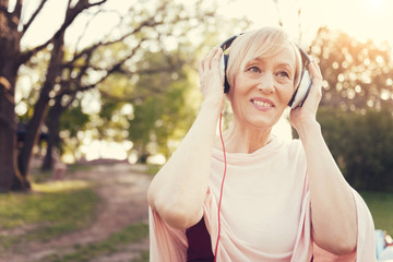 Pleasurable entertainment. Happy cheerful senior woman wearing headphones and smiling while listening to music