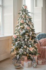 Fototapeta na wymiar A Christmas tree near the window is decorated with toys and flower in pastel colors. Christmas gifts under the Christmas tree