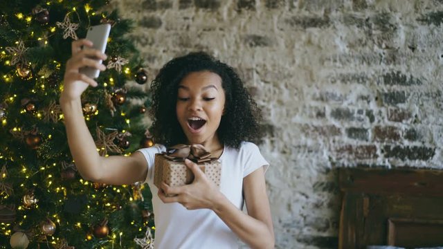 Funny mixed race girl taking selfie pictures on smartphone camera at home near Christmas tree