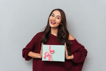Fototapeta premium Cheerful brunette girl in stylish knitted sweater holding gift box, looking at camera