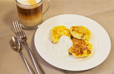 Freshly fried curd cheese cakes with sour cream and a cup of coffee with milk on the table. Homemade hot breakfast