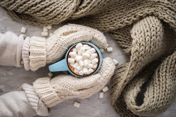 Christmas and New Year cozy holiday composition with scarf, woman hands in mittens, mugs with cocoa or chocolate, marshmallow on the gray concrete background. Flat lay, top view.