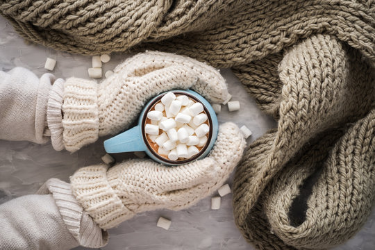 Christmas and New Year cozy holiday composition with scarf, woman hands in mittens, mugs with cocoa or chocolate, marshmallow on the gray concrete background. Flat lay, top view.
