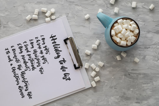 Christmas And New Year Cozy Holiday Composition With To Do List, Mug With Cocoa Or Chocolate, Marshmallow On The Gray Background. Flat Lay, Top View.