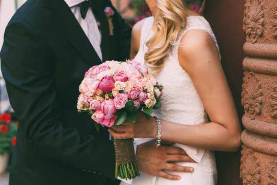 Beautiful tanned bride in a white fitting dress and groom in tuxedo are standing next to the wall and holding a wedding bouquet. Groom embrace woman by the waist. Outdoors
