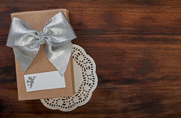 Wrapped gift box with silver ribbon bow on a dark walnut wood, on lace napkin with white card and silhouette of angel, top view, close up