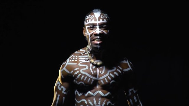 A black man in ritual patterns on his body strains and roars in the dark, slow motion