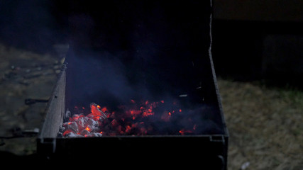 Man placing raw meat onto a hot barbecue to grill using a pair of wooden tongs, close up view over a dark background. Dinner party, barbecue and roast pork at night. Grill background
