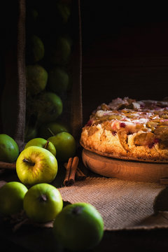 Homemade Apple pie on a round wooden board with green apples, cinnamon and red currants. Food concept. Low key image. An intimate, homely atmosphere. A beam of light on the cake and apples