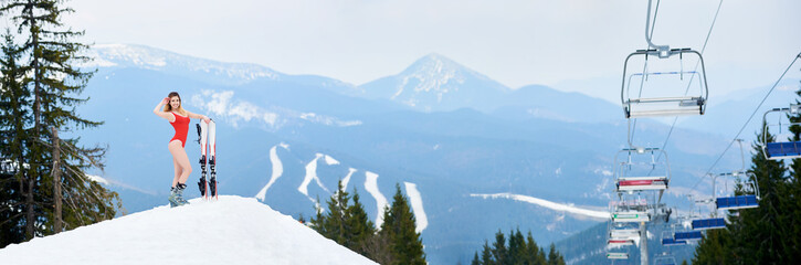 Panoramic view of winter ski resort Bukovel. Beautiful woman skier wearing red swimsuit, posing with skis on the top of the snowy slope. Mountains, forests, ski slopes and ski-lift on the background