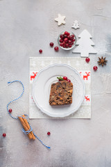 A piece of traditional Christmas English pudding (fruit cake) on white plate on a concrete light background. Christmas decor of the table. Funny napkins with reindeers Santa Claus.  Flat lay, top view