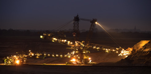 Giant excavator in an open-cast coal mine in the night, with orange and yellow glowing lights. Huge...