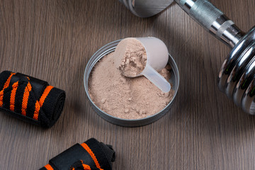 Whey protein in a measuring bucket with a metal dumbbell on a wooden background