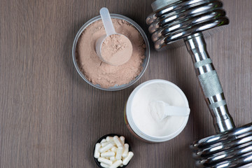 Whey protein in a measuring bucket, amino acids, sports capsules and a metal dumbbell on a wooden background copy space